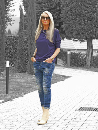 Frau trägt OFF#DLY Denim Destroy Alassio blue: schmal geschnittene, distressed Skinny-Jeans mit Used-Details. Kombiniert mit lila Oberteil, goldenen Accessoires, beige Stiefeletten und Tattoos im Freien.