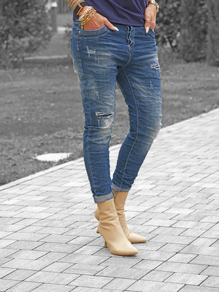 Person in schmalen, beschädigten OFF#DLY Denim Detroy Alassio blue Jeans mit umgeschlagenen Bündchen und beige Stiefeln, Hände in Taschen, trägt goldene Armbänder und Uhr.