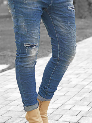 OFF#DLY Denim Detroy Alassio blue: Schmale, leicht zerstörte Jeans mit Knierissen, umgeschlagenen Säumen und komfortablem Stretch, getragen mit tanfarbenen Stiefeletten auf einem gepflasterten Weg.