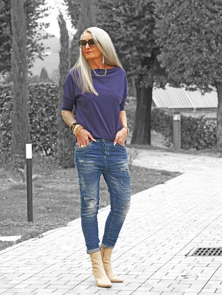 Frau trägt OFF#DLY Denim Detroy Alassio blue Jeans mit leicht zerstörtem Look und umgeschlagenen Bündchen, kombiniert mit lila Oberteil und cremefarbenen Stiefeletten, auf einem gepflasterten Weg.