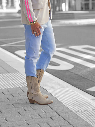 Person trägt OFF#DLY Denim Milano lightblue, eine lässige, leicht zerstörte Jeans. Kombiniert mit Cowboy-Stiefeln und gestreifter Jacke. Jeans zeigt klassischen Five-Pocket-Stil und bequemen Sitz.