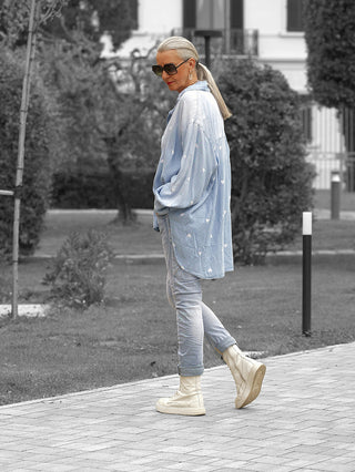 Frau trägt die OFF#DLY Denim Bluse Pia lightblue mit weißen Herzmustern, oversized und lässig über schmalen Jeans, kombiniert mit cremefarbenen Stiefeln und Sonnenbrille beim Spaziergang im Park.