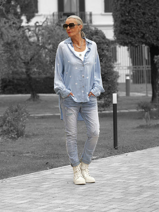 Frau in OFF#DLY Denim Bluse Pia lightblue mit Herzmuster, über weißem Tanktop, kombiniert mit Jeans und weißen Sneakern, in einem lässigen, modernen Look auf einem gepflasterten Weg.