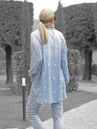 Person von hinten in einem formalen Garten, trägt die OFF#DLY Denim Bluse Pia lightblue mit Herzmuster, oversized Schnitt und tiefem Pferdeschwanz, kombiniert mit passender Jeans und Sonnenbrille.