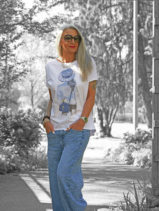 Frau in oversized weißem OFF#DLY Tee Hat mit Hut-Print, locker sitzende blaue Jeans und auffälligem Schmuck, auf einem schattigen Weg stehend.