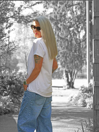 Person mit langen blonden Haaren trägt ein OFF#DLY Tee Hat white/blue, oversized, mit auffälligem Fashion-Print. Sonnenbrille, Jeans und Tattoos sichtbar, in sonnigem Gartenweg stehend.