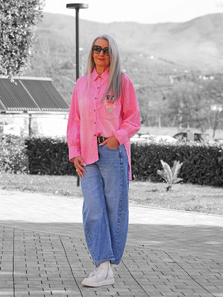 Mature Frau trägt die OFF#DLY Bluse Anna pink im Oversize-Stil mit Destroy-Look und Brusttasche, kombiniert mit lässigen blauen Jeans und weißen Sneakers.