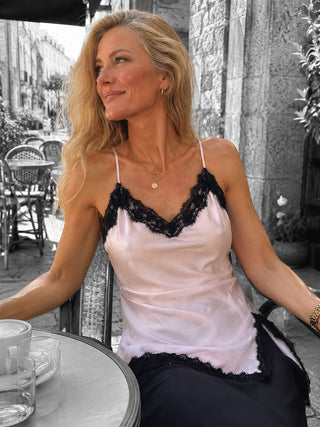 Frau in einem Outdoor-Café, trägt das OFF#DLY Top Spitze Veronika rosa/black, ein elegantes Satin-Top mit schwarzer Spitze, kombiniert mit goldenen Halsketten und kleinen Ohrringen.