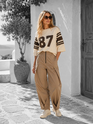 Frau trägt Co Couture Trice Pleat Pant in Sand, eine beige, weit geschnittene Hose mit elastischem Bund und dezenten Falten, kombiniert mit einem lockeren Sweatshirt und Sneakers.