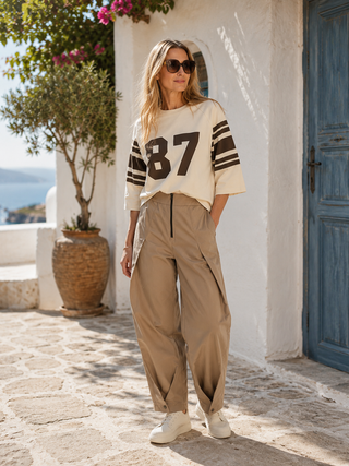Frau in Co Couture Ely 87 Tee Sweat brown/bone mit „87“-Print und Streifen, kombiniert mit weiten Khaki-Hosen, steht vor einer mediterranen Kulisse mit blauer Tür und Olivenbaum.
