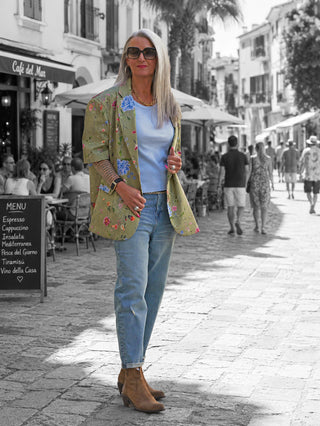Frau in Lumina Blazer Daphne kiwi mit floralem Print, kombiniert mit hochtaillierten Jeans und braunen Stiefeletten, steht auf Kopfsteinpflaster vor Café.