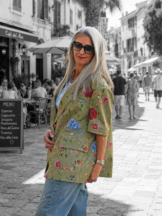 Frau trägt den Lumina Blazer Daphne kiwi mit floralem Print und Oversize-Schnitt auf einer sonnigen Straße, kombiniert mit Jeans und Sonnenbrille, vor einem Café.