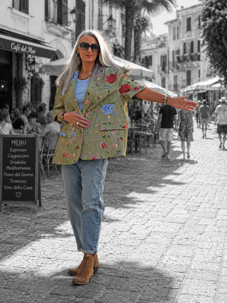 Frau trägt Lumina Blazer Daphne kiwi mit floralen Stickereien, kombiniert mit Jeans und Stiefeletten, auf einer belebten Straße. Der Blazer hat einen oversize Schnitt und kurze Ärmel.