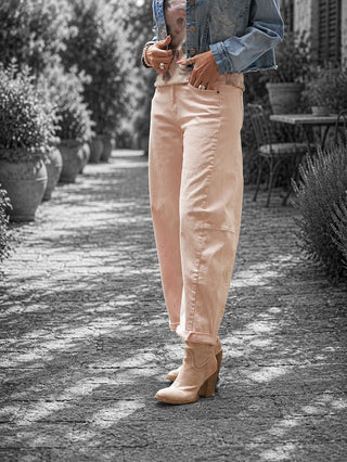Person trägt OFF#DLY Pant Ronja rosa, eine locker geschnittene, am Knöchel gekrempelte Hose. Kombiniert mit einer Jeansjacke und tanfarbenen, heeled Stiefeletten auf einer gepflasterten Straße.