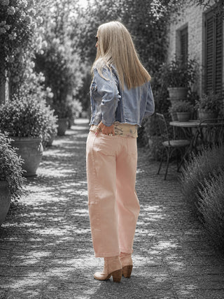 Frau in Gartenhof trägt OFF#DLY Pant Ronja rosa, eine hochgeschnittene, weit geschnittene Hose in sanftem Rosé, kombiniert mit einem blauen Jeansjacke und hellen Stiefeletten.