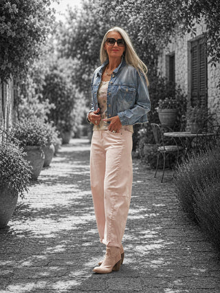 Ältere Frau trägt die OFF#DLY Pant Ronja rosa mit modernem Barrel-Fit und sanfter rosé Farbe, kombiniert mit einem Denim-Jacke und beigen Stiefeletten auf einem sonnigen Steinweg.
