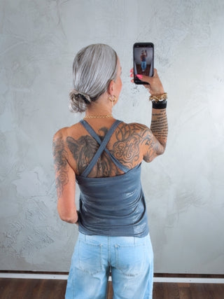 Person mit hellgrauem Dutt trägt das Cotton Candy Tank Top Lois blue ash mit gekreuzten Trägern, kombiniert mit weiten Jeans. Beide Arme und oberer Rücken sind mit Tattoos bedeckt.