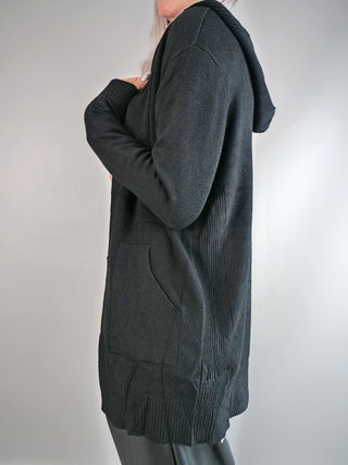 Person trägt die OFF#DLY Strickjacke mit Kapuze schwarz, eine lange, schwarze Strickjacke mit Rippenbündchen und aufgesetzten Taschen, kombiniert mit schwarzen Hosen oder Leggings.