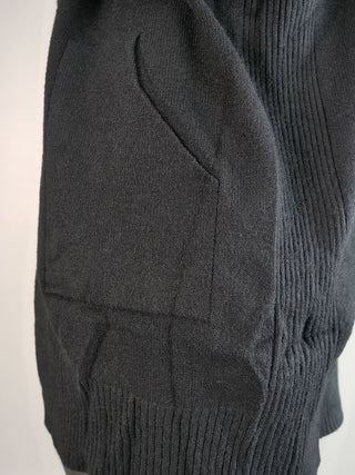 Nahaufnahme der OFF#DLY Strickjacke mit Kapuze schwarz, zeigt eine geschwungene Seitentasche, breites Rippband unten und vertikale Rippstrickmuster, die für stilvolle Texturkontraste sorgen.