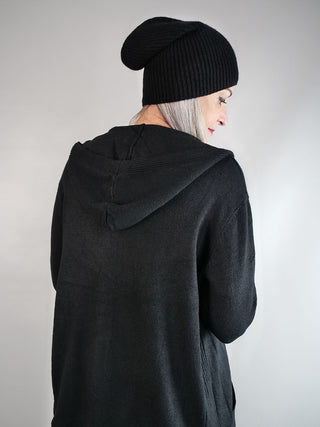 Person von hinten in einer langen, schwarzen OFF#DLY Strickjacke mit Kapuze und schwarzer Mütze, leichte graue Haare sichtbar, vor hellem Hintergrund.