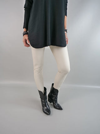 Eine Frau trägt die OFF#DLY Legging Creme, kombiniert mit einem schwarzen Shirt und schwarzen Stiefeln. Die cremefarbene Legging aus 100% Polyester bietet einen eleganten, modernen Look.