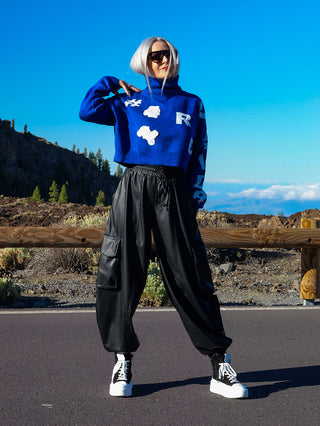 Frau trägt #VDR Wide Leg Pant aus Fake-Leder, kombiniert mit einem blauen Pullover, zu sehen in einer Outdoor-Szene. Nahe der Füße sind Schuhe erkennbar.