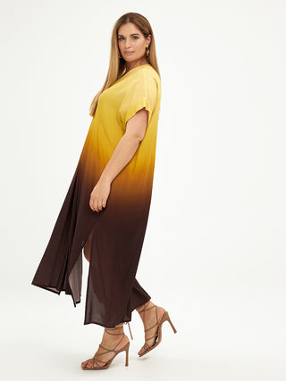 Frau trägt Mat Dress 8101.7078 in blassem Gelb, aus 100% Viskose, mit auffälligem gelb-braunem Muster. Nahaufnahme zeigt Füße, betont den fließenden Fall des Kleides.