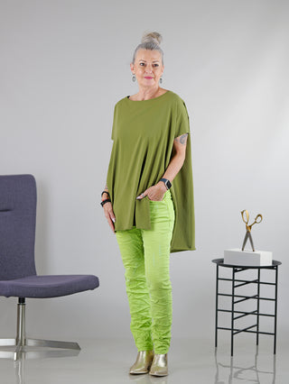 Frau trägt SOHO-t T-Shirt Mogano khaki, lässige Tunika mit seitlichem Schlitz und Rundhals, hinten länger geschnitten. Sie steht in einem Raum, kombiniert mit grüner Hose und Armbanduhr.