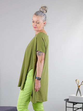 Frau trägt SOHO-t T-Shirt Mogano khaki, lässige Tunika mit seitlichem Schlitz, hinten länger geschnitten. Rundhals-Design. Sichtbarer grüner Stoff und Arm-Tattoo. Nahe Umgebung mit Schere und Uhr.