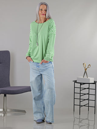 Frau trägt das OFF#DLY Ringelshirt in Grün-Weiß mit langen Ärmeln und Rundhalsausschnitt, locker geschnitten, kombiniert mit blauen Jeans, Hände in den Taschen, lässig und stilvoll.