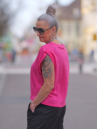 LTC Tee Muflier fuchsia aus Leinen, getragen von einer Frau mit tätowiertem Arm, grauen Haaren und Sonnenbrille. Oversize-Schnitt mit angeschnittenen Ärmeln und Cerises-Schriftzug aus Strasssteinen auf dem Rücken.