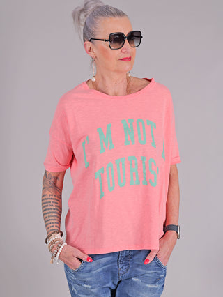 Frau trägt das One Tee Margareth coral mit blauem Aufdruck, kombiniert mit Jeans. Sie trägt Sonnenbrillen und Armband. Kurzarm-T-Shirt aus 100% Baumwolle, ideal für einen lässigen Look.