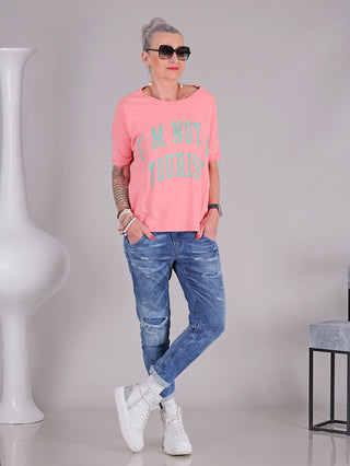 Eine Frau trägt das One Tee Margareth coral mit kurzem Arm und Print vorne und hinten, kombiniert mit blauen Jeans und weißen Schuhen.