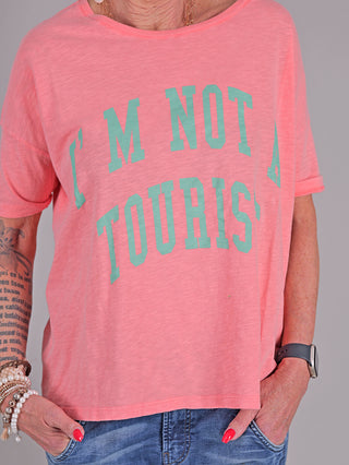 One Tee Margareth coral, getragen von einer Person, zeigt einen lässigen Kurzarmstil mit auffälligem Front- und Rückendruck. Das T-Shirt wird mit Jeans kombiniert und ergänzt durch ein Armband und eine Uhr.