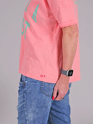 Person trägt das One Tee Margareth coral, ein Kurzarmshirt mit auffälligem Print vorne und hinten, kombiniert mit Jeans. Sichtbar ist auch eine Uhr am Handgelenk.