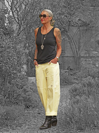 Ältere Frau trägt OFF#DLY Pant Sia yellow, eine locker sitzende, leicht verkürzte Hose mit elastischem Bund und dezenten Nahtdetails, kombiniert mit einem schwarzen Tanktop und schwarzen Stiefeln.