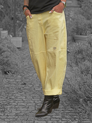 Person trägt OFF#DLY Pant Sia yellow, eine locker geschnittene Hose mit umgeschlagenem Saum, kombiniert mit schwarzen Stiefeletten. Hände in Taschen, betont durch Armbänder und Tätowierungen.