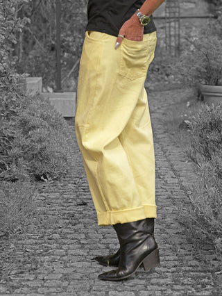 Person in knöchellanger OFF#DLY Pant Sia yellow mit lockerer Passform und umgeschlagenem Saum, dazu schwarze Lederstiefeletten. Die rechte Hand steckt lässig in der Hosentasche.