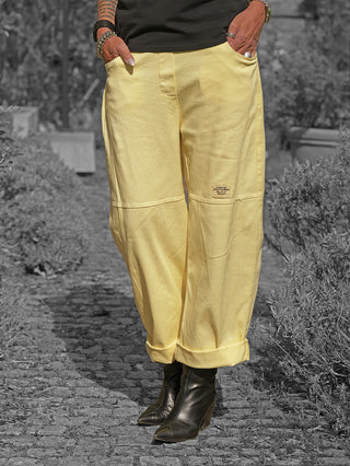 Person trägt OFF#DLY Pant Sia yellow, eine locker geschnittene, leicht verkürzte Hose mit umgeschlagenen Bündchen, kombiniert mit schwarzen Lederstiefeletten. Hände in den Taschen, mehrere Ringe und Armbänder sichtbar.