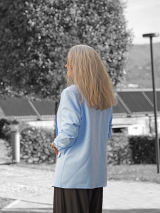 Person von hinten in einem OFF#DLY Blazer Dana lightblue, mit lässigem Schnitt, Reverskragen und aufgesetzten Taschen, stehend auf einem gepflasterten Weg mit Pflanzen und Gebäude im Hintergrund.
