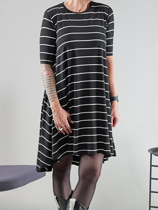 Frau trägt SOHO-t Kleid Anna schwarz/weiß, gestreift mit asymmetrischem Schnitt. Sichtbar sind schwarze Stiefel und eine Handtasche. Modelgröße: 172 cm, trägt Größe XS.