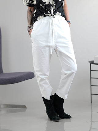 Person trägt Thom Krom Pant in Off-White, lässige Jogginghose mit Taschen, Gummibund und Kordelzug, aus Baumwoll-Elastan-Mix. Die Hose fällt größer aus und bietet einen entspannten, modischen Look.