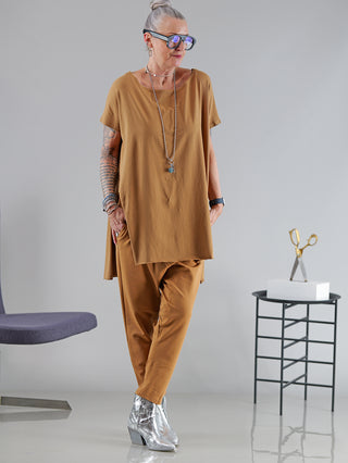 Frau in braunem SOHO-t T-Shirt Mogano zimt, lässige Tunika mit seitlichem Schlitz und längerem Rückenteil, vor einem Stuhl.